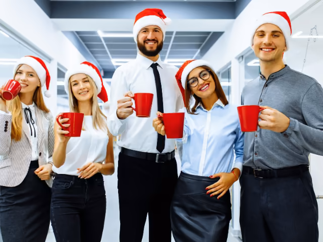 Equipo de trabajo con gorros navideños brindando con pocillos rojos en la oficina antes del evento de fin de año