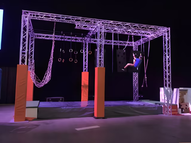 Estructura truss de aluminio armada como cubo autoportante, con anillos, red de cuerda y muro que una persona escala en una actividad de evento empresarial