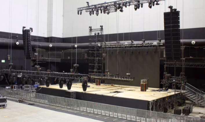 Montaje de gran formato en un recinto amplio, con tarima, estructura truss con iluminación colgada y arreglos de sonido a los costados