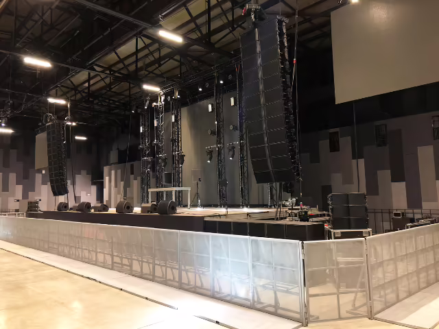 Line array colgado a lado y lado de la tarima durante el montaje de un evento empresarial en un pabellón cubierto