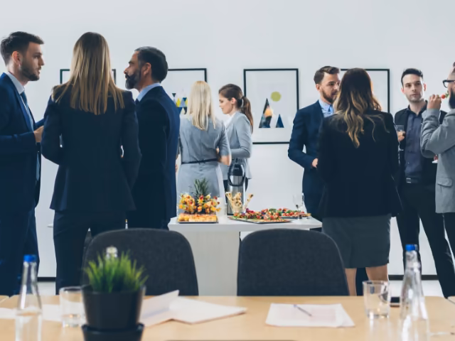 Colaboradores de traje conversando alrededor de una mesa de pasabocas durante un evento empresarial en la oficina